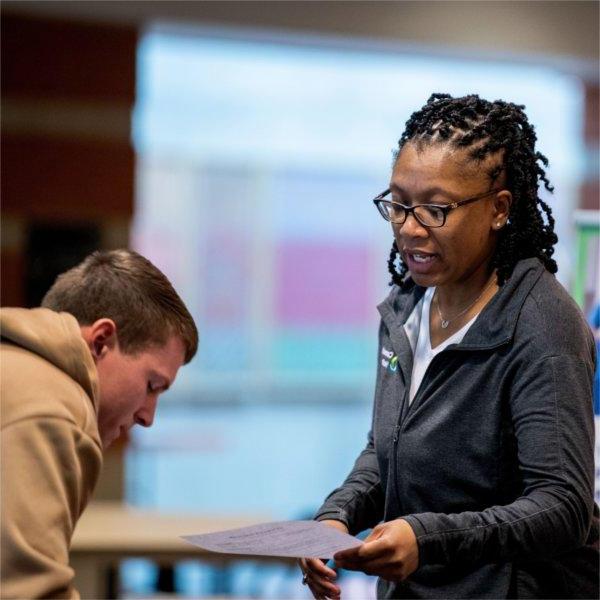 A person holding a paper speaks to a person who is bending down toward the table.