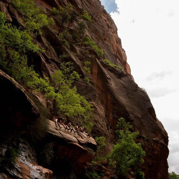 People sit on the edge of a cliff with their legs dangling over. The rock formation continues up and behind them and is covered with dotted greenery.