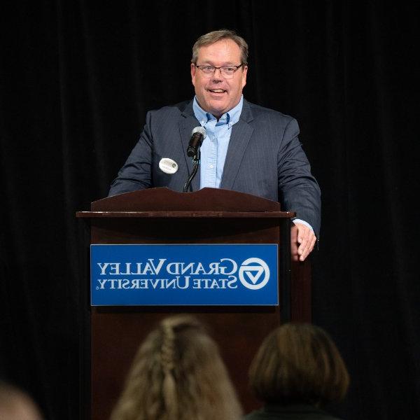 Greg Warsen at podium in blue suit coat and light blue shirt with no tie; podium has GVSU wordmark on it