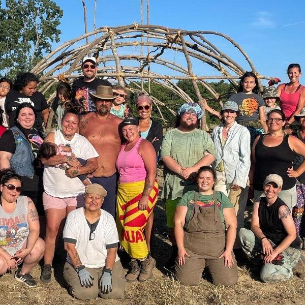 large group of people standing in front a teaching lodge, which is a sacred space to Native Americans constructed from saplings.